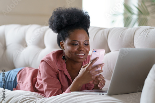 Smiling African American millennial woman with curly afro hairstyle in pink shirt lying on couch at home, using cellphone and chatting in social networks, working on laptop. 
