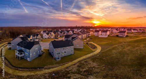 Aerial view of single family homes covered with blue, white, beige vinyl or brick façade surrounding dead end neighborhood street, American residential community dramatic colorful, yellow, orange sky