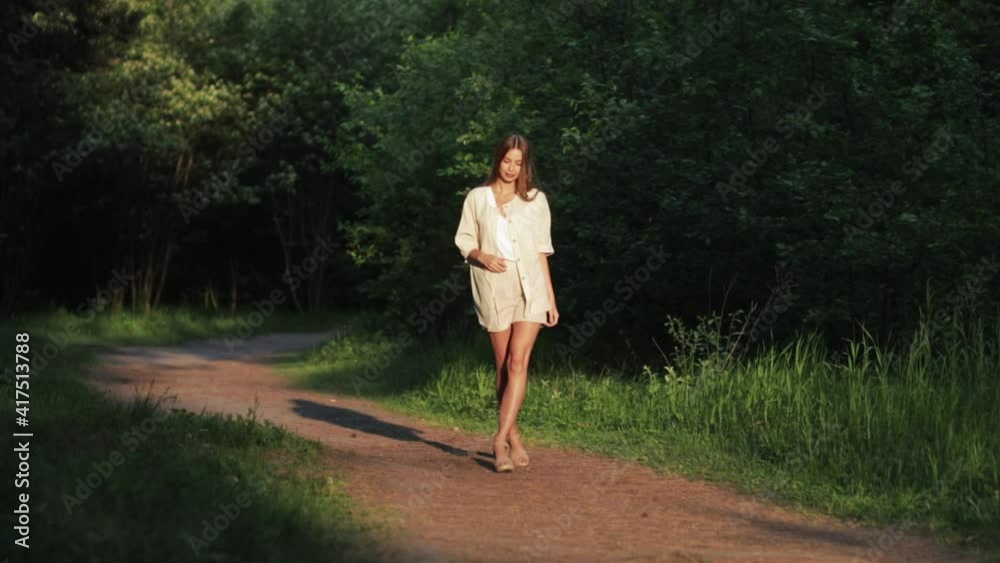 A beautiful woman smiles as she walks along a path surrounded by foliage and trees. Front view of a woman in a dress walking in a summer Park. Slow motion A beautiful woman smiles as she walks along a path surrounded by foliage and trees. Front view of a woman in a dress walking in a summer Park. Slow motion