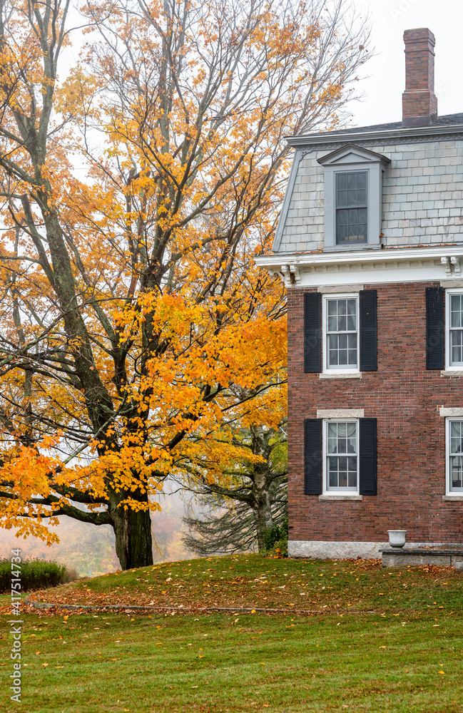 An old brick house with an attic and a chimney stands on a hill next to an autumn yellow maple tree in New England in foggy weather An old brick house with an attic and a chimney stands on a hill next to an autumn yellow maple tree in New England in foggy weather