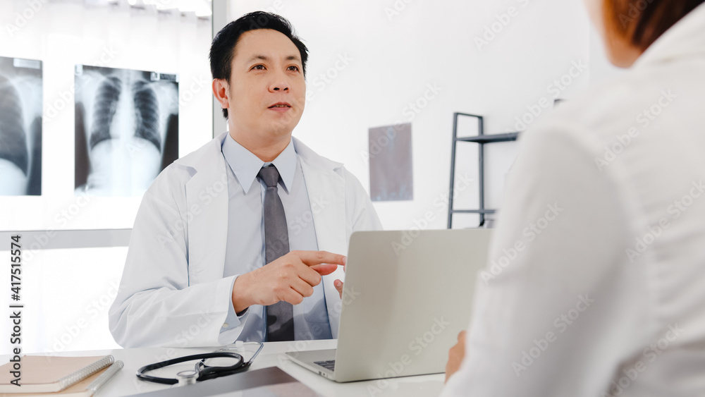 Serious Asia male doctor in white medical uniform using computer laptop is delivering great news talk discuss results or symptoms with female patient sit at desk in health clinic or hospital office.
