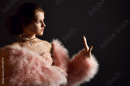 Portrait of kid girl, young lady in evening strapless dress and fur coat with big sleeves holding hand up, gesturing stop, enough sign or touching something and looking aside over dark background photo