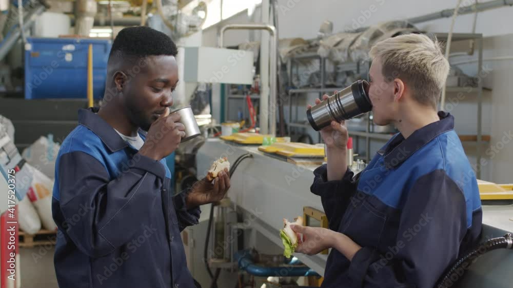 Medium shot of cheerful male and female factory workers in uniforms laughing and chatting while enjoying their sandwiches during lunch break in workplace Medium shot of cheerful male and female factory workers in uniforms laughing and chatting while enjoying their sandwiches during lunch break in workplace