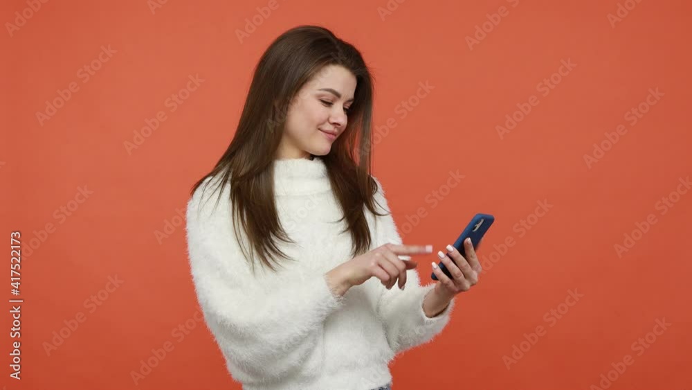 Profile portrait smiling brunette woman in fluffy white sweater swiping, scrolling and typing on smartphone with smile on her face, using dating app. Indoor studio shot isolated on orange background Profile portrait smiling brunette woman in fluffy white sweater swiping, scrolling and typing on smartphone with smile on her face, using dating app. Indoor studio shot isolated on orange background