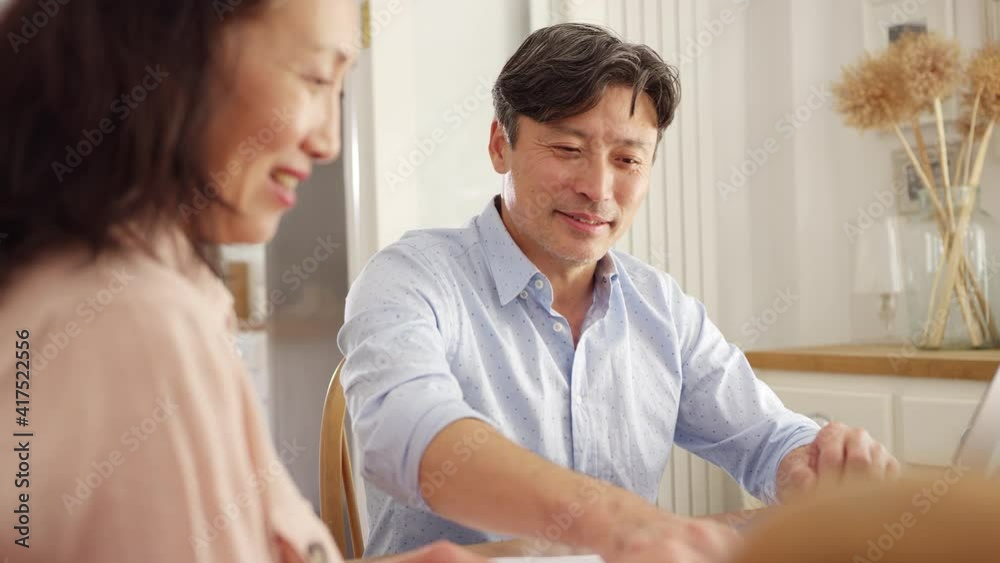 Mature Asian couple sitting at table at home using laptop to organise household bills and finances - shot in slow motion Mature Asian couple sitting at table at home using laptop to organise household bills and finances - shot in slow motion