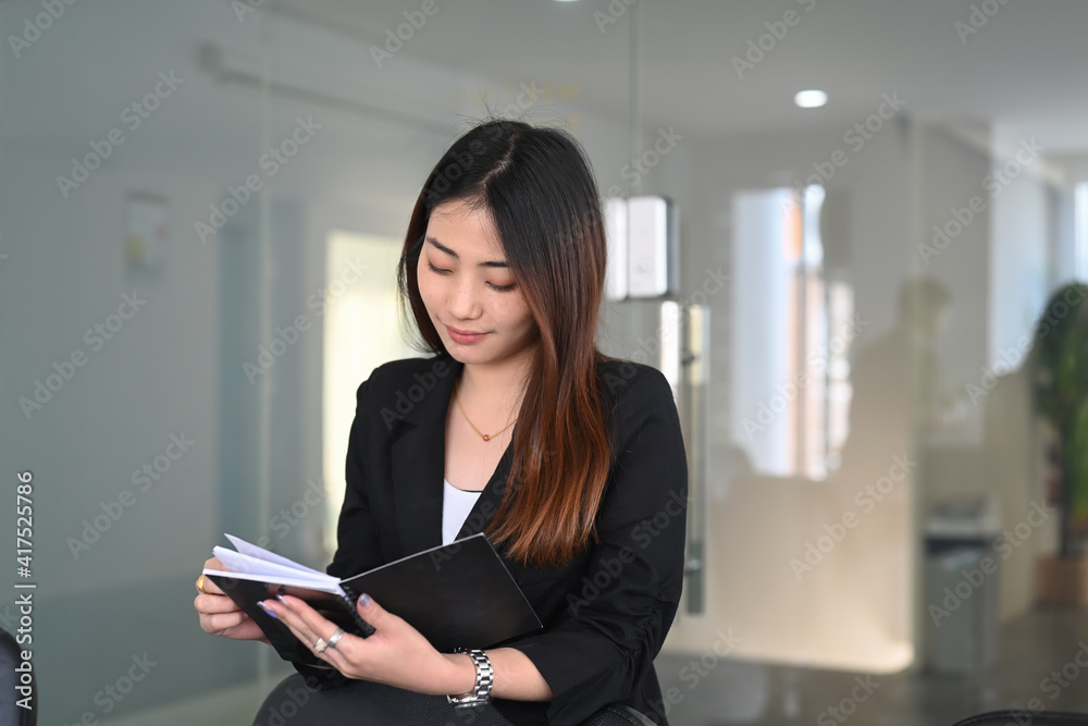 Smiling businesswoman reading information on notebook while standing in office. Smiling businesswoman reading information on notebook while standing in office.