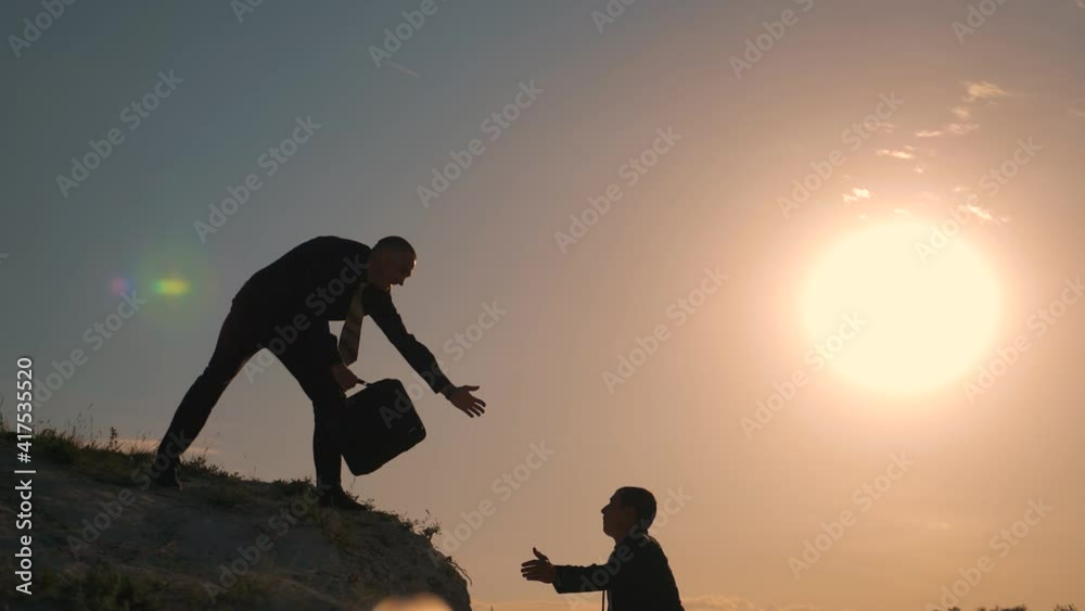 Businessmen climb to the top of the mountain, careers and one of them gives a helping hand. Silhouette of two men at sunset. Dressers help in crisis, overcoming obstacles. The concept of teamwork.