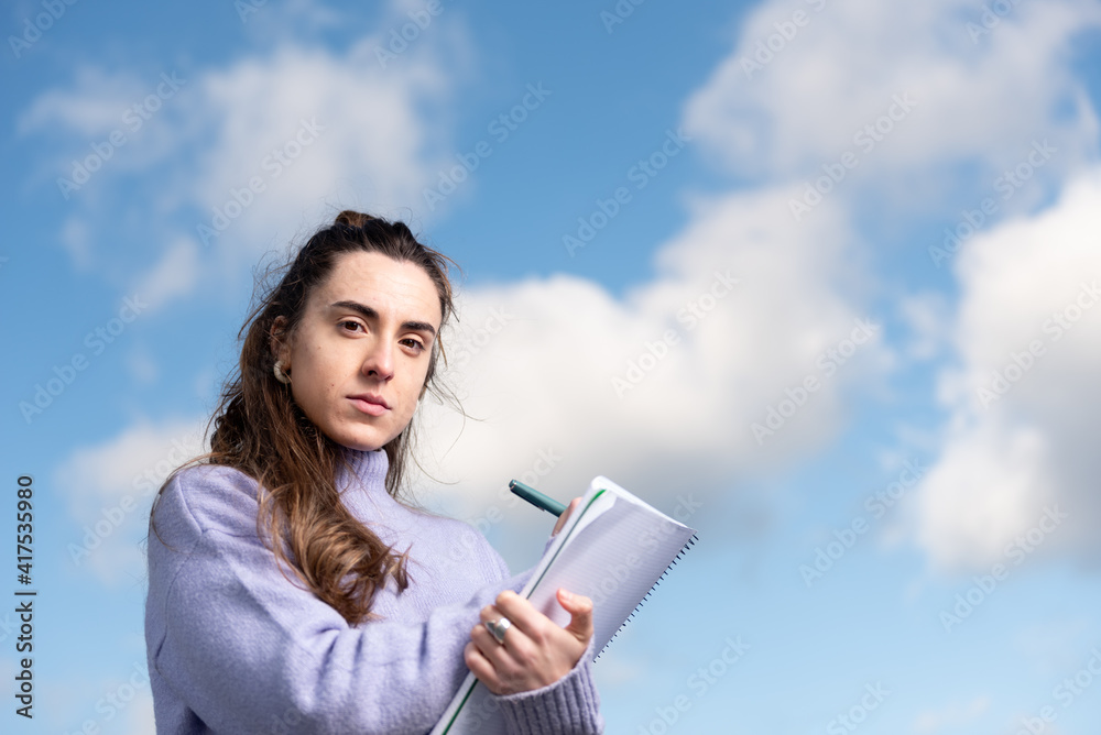 Closeup portrait of a young pretty woman writting on a notebook while looking with serious face to the camera with a sky full of clouds as background. Closeup portrait of a young pretty woman writting on a notebook while looking with serious face to the camera with a sky full of clouds as background.