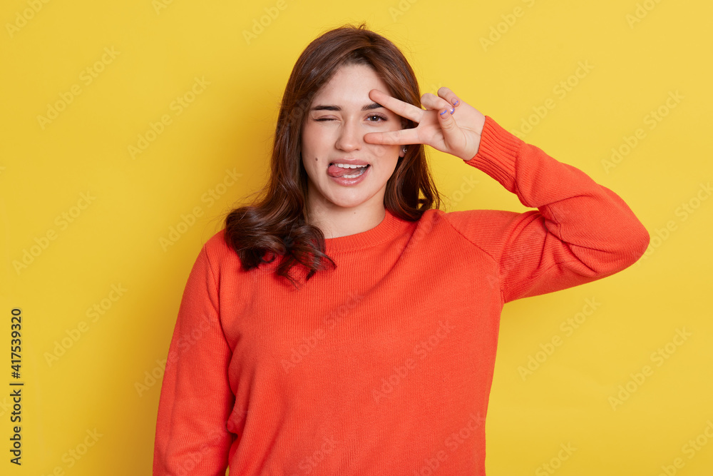 European dark haired lady with funny expression, posing isolated over yellow background, showing tongue, frowning face, winking one eye and cover other one with v sign, victory gesture. European dark haired lady with funny expression, posing isolated over yellow background, showing tongue, frowning face, winking one eye and cover other one with v sign, victory gesture.