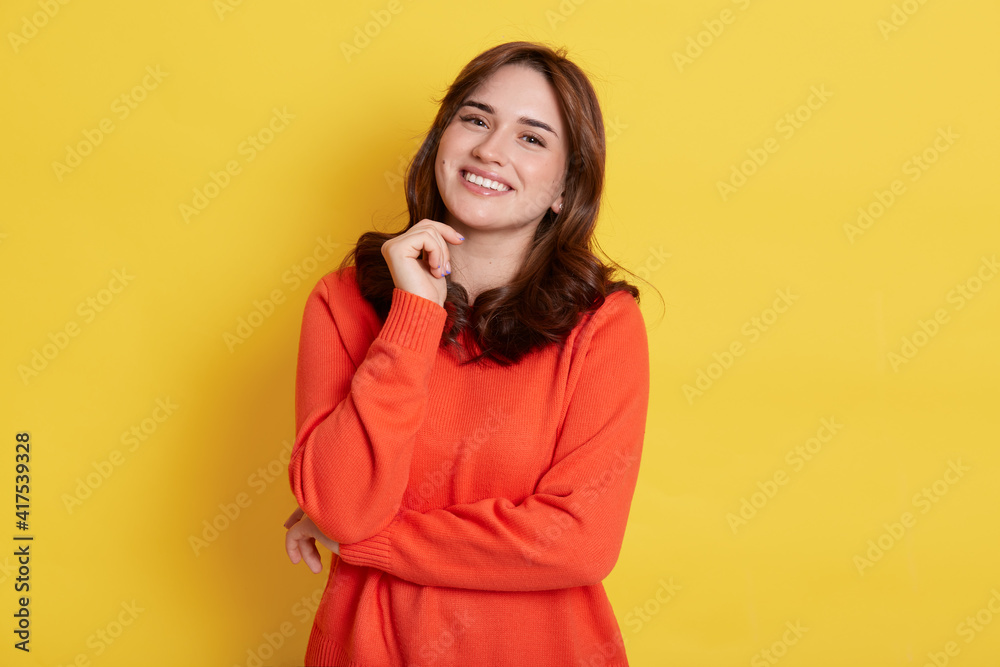 Happy smiling dark haired woman with pleasant appearance, wearing casually, looking at camera with toothy smile, keeping fist under chin, standing isolated over yellow background. Happy smiling dark haired woman with pleasant appearance, wearing casually, looking at camera with toothy smile, keeping fist under chin, standing isolated over yellow background.
