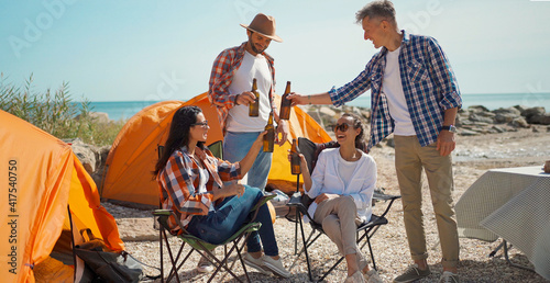 four adult friends travelers enjoying cold beer at hot sunny day in camping on sea coast. joyful friends on picnic drinking cold beverages, girls relaxing in camp chairs