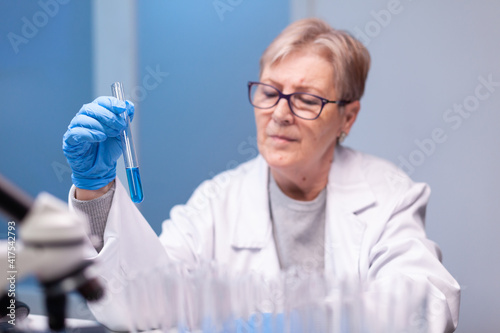 Scientist senior woman looking into test tube for biochemistry test. Doctor in white coat research a new experiment in laboratory, analyzing biotechnology work with modern equipment