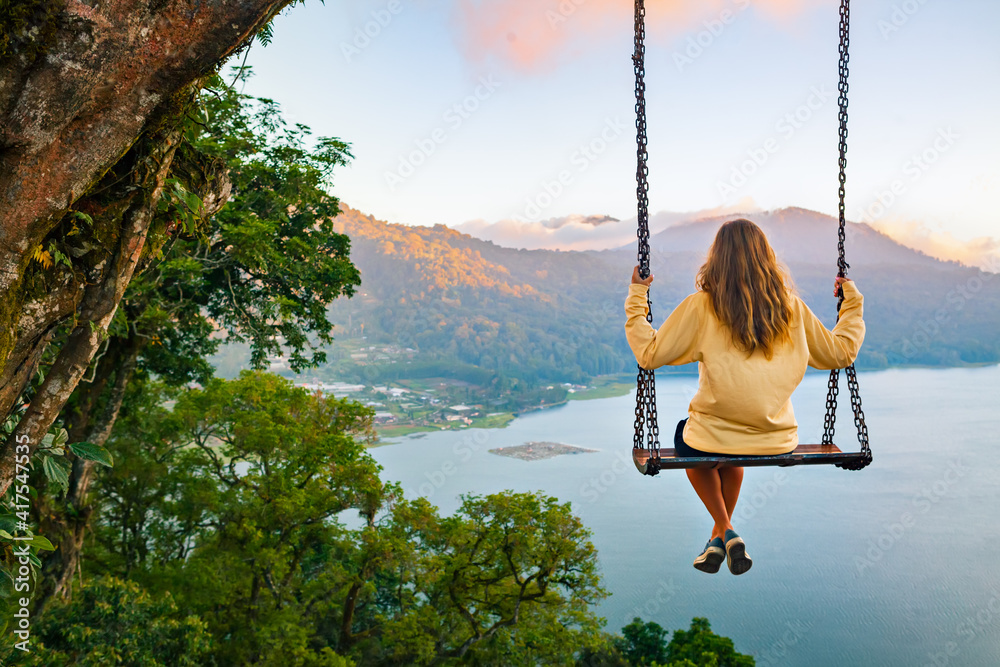 Summer vacation. Young woman sit on tree rope swing on high cliff above tropical lake. Happy girl looking at amazing jungle view. Buyan lake is popular travel destinations in Bali island, Indonesia - obrazy, fototapety, plakaty 
