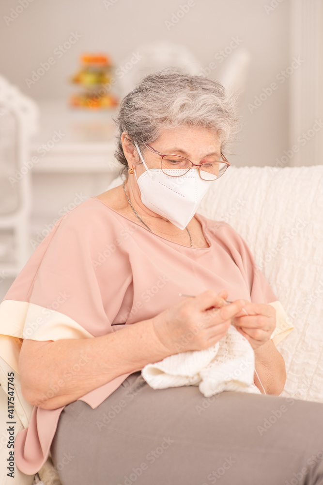 A pretty retired old woman with a medical mask knits at home during a coronavirus lockdown. Ways not to get bored during lockdown and develop while stay at home. Quarantine during coronavirus pandemic
