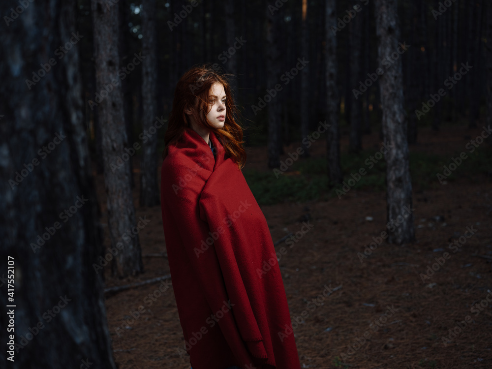 A red-haired woman with a plaid on her shoulders walks in the forest near the trees