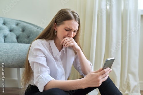 Sad crying young woman with smartphone sitting on the floor at home