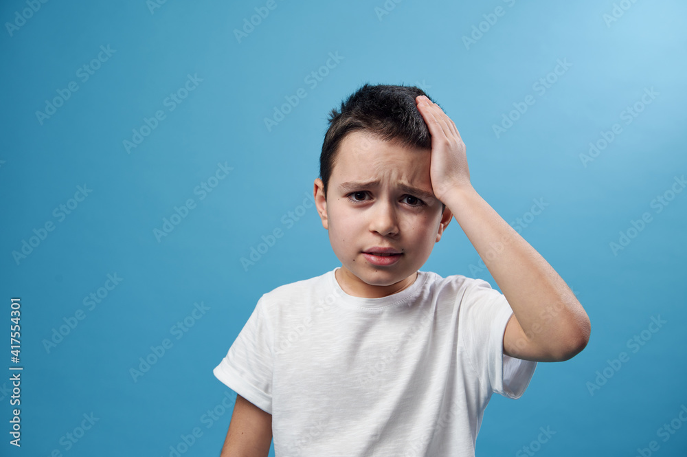 Boy holding his temple. Facial emotion on blue background with copy space Boy holding his temple. Facial emotion on blue background with copy space