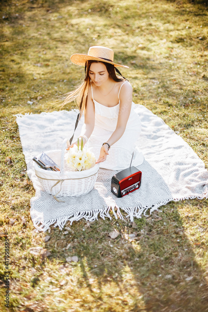 Obraz premium Girl with a straw hat in the spring in the park. Brunette with long hair sitting on a plaid on a background of summer nature. Youth and beauty.