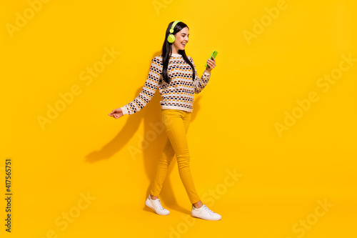Full size profile side photo of young beautiful happy excited girl in headphones go walk use phone isolated on yellow color background