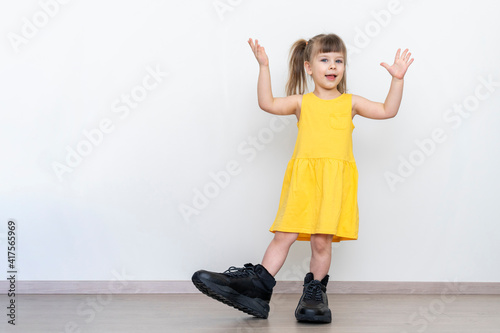 playful little girl in a yellow dress posing in huge mens boots against a gray wall, mock up with copy space