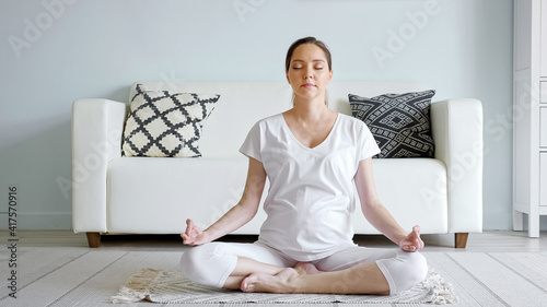 Tranquil young pregnant woman in white meditates sitting in lotus pose on floor rug near designer sofa in spacious room at home close-up