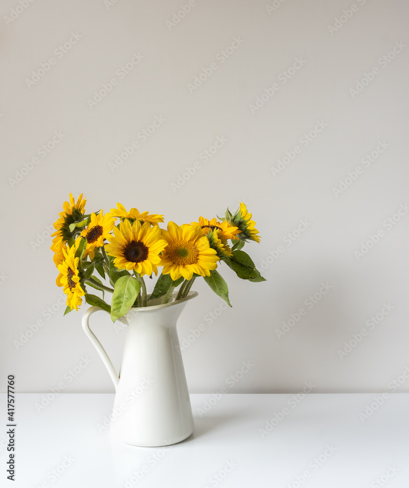 Vertical close up of sunflowers in white jug on shelf against neutral wall background (selective focus)