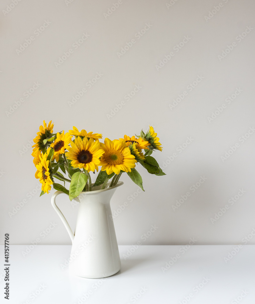 Vertical close up of sunflowers in white jug on shelf against neutral wall background (selective focus) Vertical close up of sunflowers in white jug on shelf against neutral wall background (selective focus)