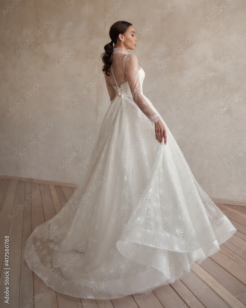 Full length bride portrait. Elegant woman with pony tail in white wedding dress spinning in beige interior. Dynamic shot. Elegant female model standing and posing in ball gown Full length bride portrait. Elegant woman with pony tail in white wedding dress spinning in beige interior. Dynamic shot. Elegant female model standing and posing in ball gown