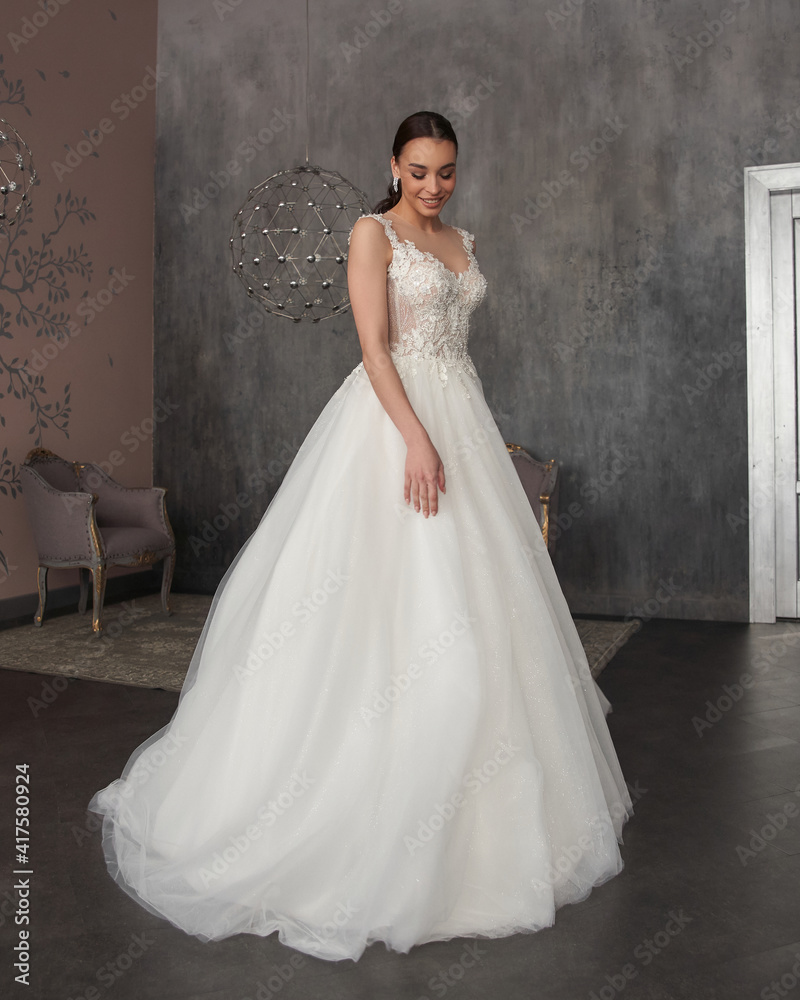 Pretty young caucasian brunette bride with make-up and pony tail hairstyle in long wedding ball gown dress standing and posing in grey interior Pretty young caucasian brunette bride with make-up and pony tail hairstyle in long wedding ball gown dress standing and posing in grey interior