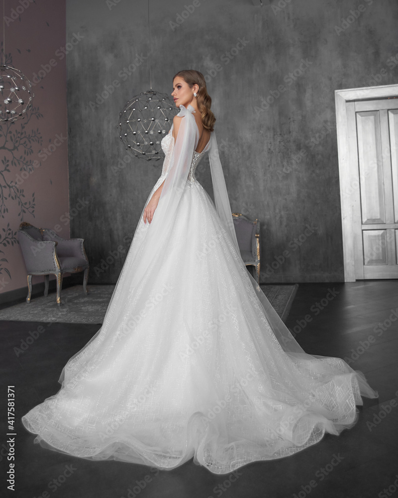 Fototapeta premium Full length bride portrait. Elegant woman with hairstyle in white wedding dress spinning in grey interior. Dynamic shot. Elegant female model standing and posing in ball gown