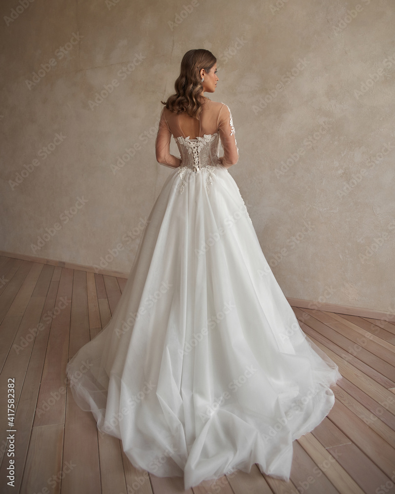 Full length portrait of young beautiful woman wearing white wedding dress. Elegant bride standing and posing against beige wall Full length portrait of young beautiful woman wearing white wedding dress. Elegant bride standing and posing against beige wall