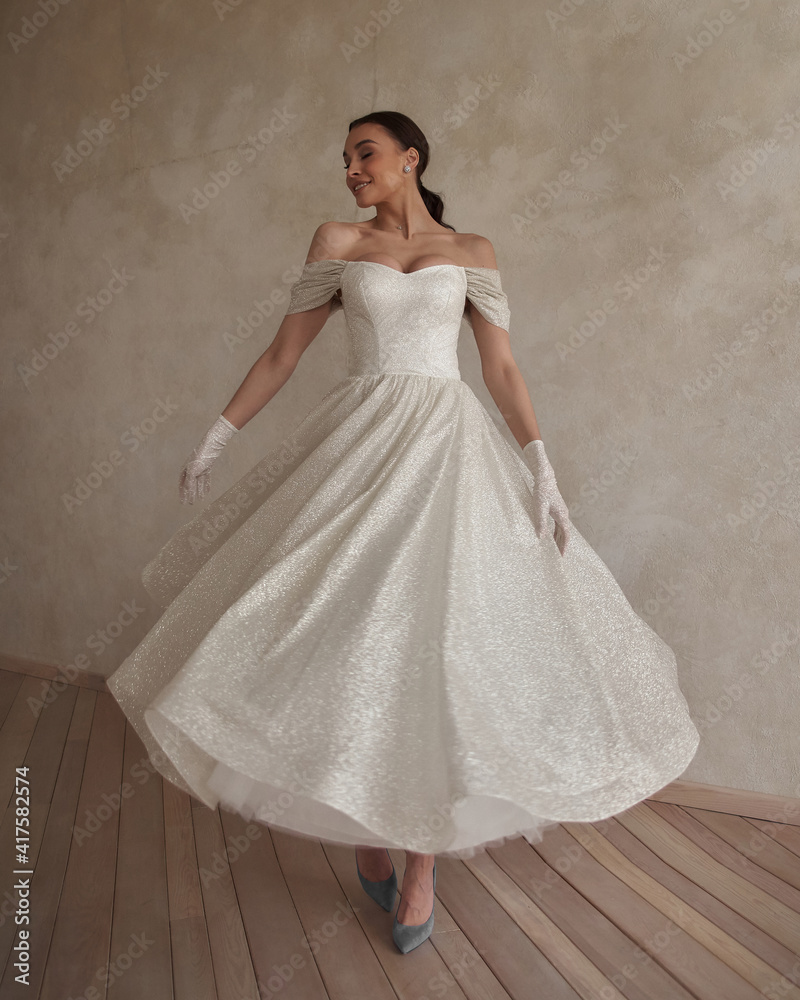 Full length bride portrait. Elegant woman with pony tail in white wedding dress spinning in beige interior. Dynamic shot. Elegant female model standing and posing in ball gown Full length bride portrait. Elegant woman with pony tail in white wedding dress spinning in beige interior. Dynamic shot. Elegant female model standing and posing in ball gown