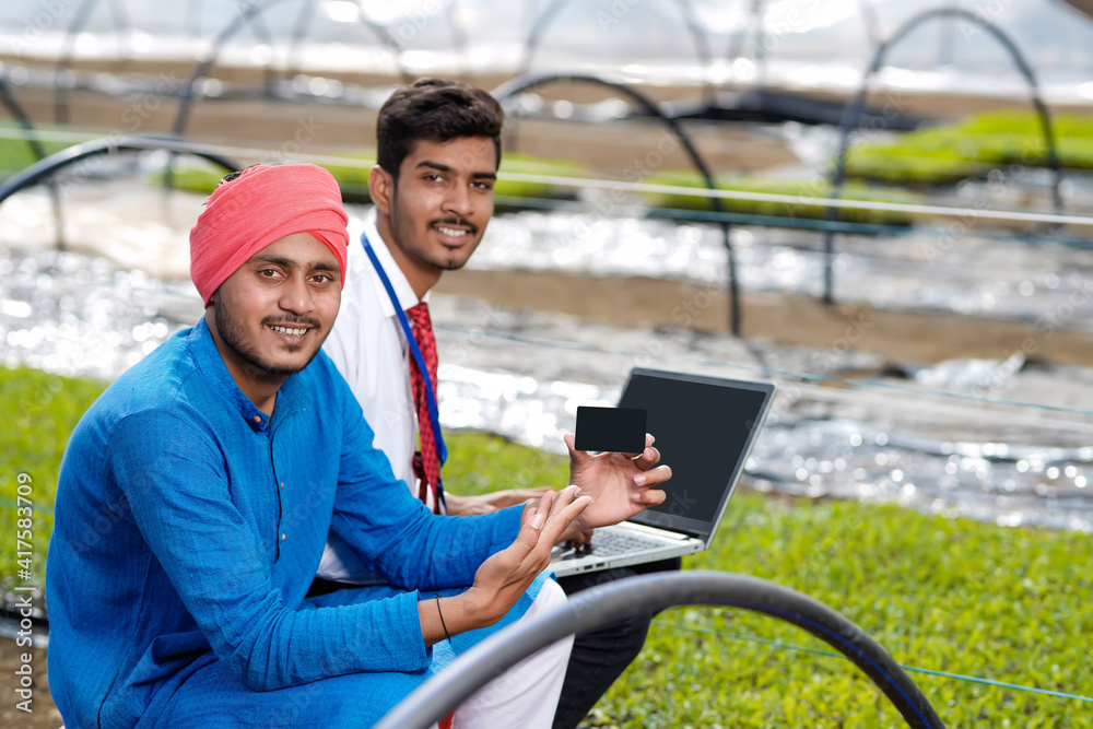Young indian agronomist showing some information to farmer in laptop at greenhouse