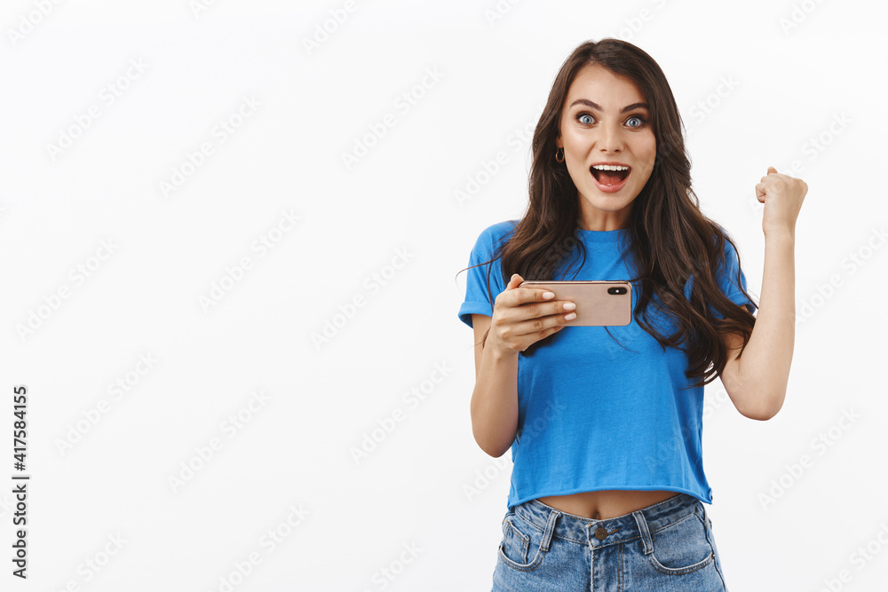 Hooray victory. Smiling happy girl compete in online challenge, watching online tennis stream on smartphone app, fist pump triumphing, smiling excited, winning in game, white background Hooray victory. Smiling happy girl compete in online challenge, watching online tennis stream on smartphone app, fist pump triumphing, smiling excited, winning in game, white background