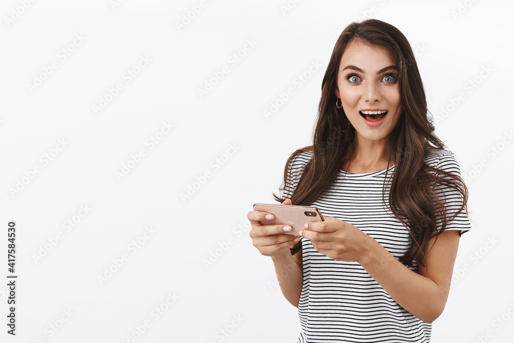 Excited and inspired good-looking caucasian female, playing awesome new game, holding smartphone horizontally, download interesting arcade, pass level, smiling camera amused, white background Excited and inspired good-looking caucasian female, playing awesome new game, holding smartphone horizontally, download interesting arcade, pass level, smiling camera amused, white background