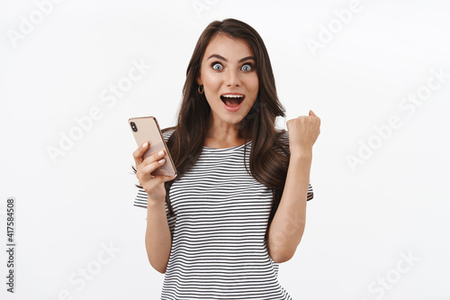 Congratulations, mobile concept. Satisfied happy and cheerful brunette female fist pump rejoicing, saying hooray or yes, reading wonderful news from message, holding smartphone, white background
