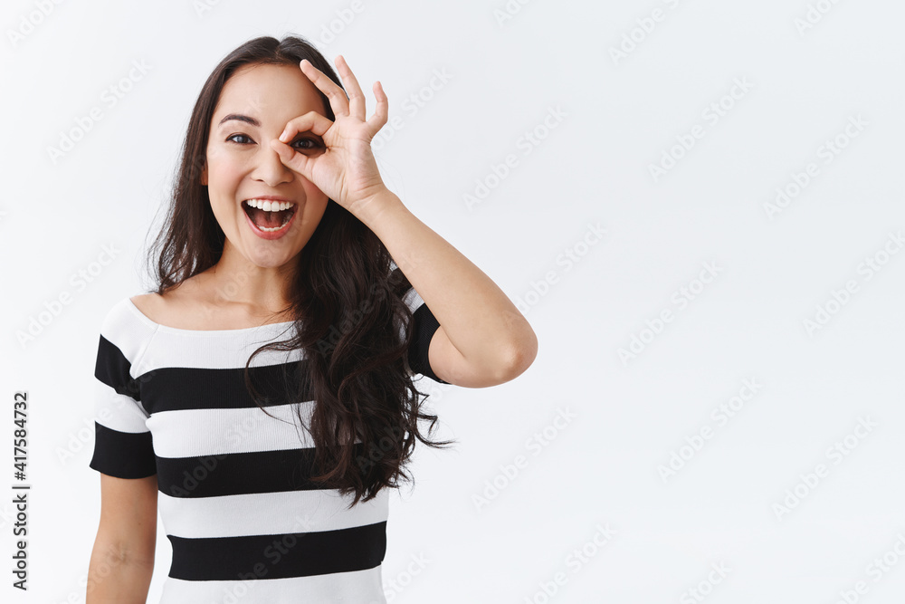 Enthusiastic happy young east-asian woman in striped t-shirt, looking through OK sign with astonishment and joy, staring entertained at wonderful event, standing white background