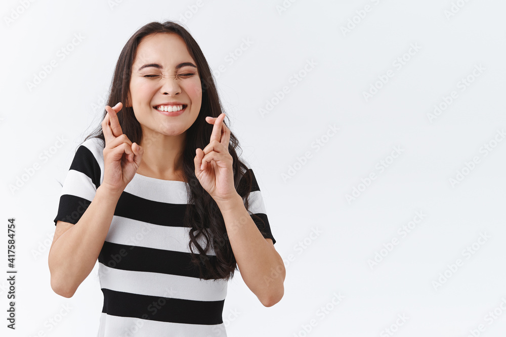 Enthusiastic, hopeful young girl keep faith, feeling lucky and amused as awaiting something important, begging for good news, keep fingers crossed, smiling with closed eyes, praying or make wish