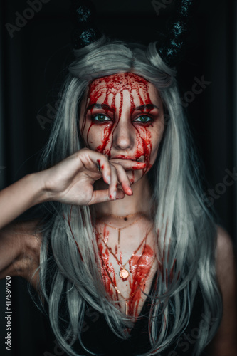 A beautiful demon girl with blood running down her face licks the blood from her hand. On the head of the horns, on the face of a beautiful makeup 