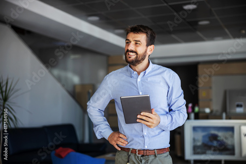 Smiling successful bearded chief standing in lobby of shipping firm, holding tablet and holding hand on hip. He is satisfied because business is going well. photo