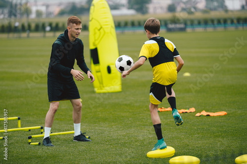 Fototapeta Naklejka Na Ścianę i Meble -  Young Man Coaching Soccer Boy. Coach on Training Session With Teenage Boy. Soccer Player Kicking Ball Standing on Stability Cushion on Grass Field. Football Player Improving Skills With Trainer