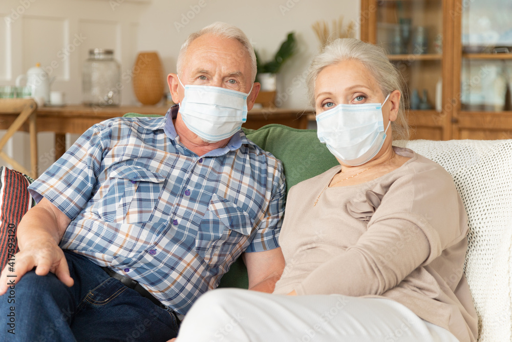 Senior couple in medical protective masks from coronavirus pandemic spread sitting on couch. Elderly man and female in face covers from covid-19 virus, staying at home. Healthcare and epidemic concept