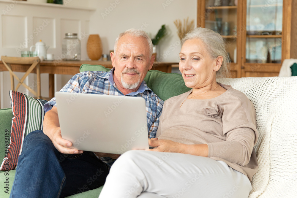 Pretty elderly gray haired couple rest on cozy couch in living room with laptop watching movie or shopping online smiling and enjoy free time together. Old generation and modern wireless technology.