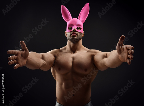 Portrait of naked handsome muscular man, athlete with perfect built body wearing pink rabbit mask stands topless and with arms outstretched towards camera, going to hug grab over dark background