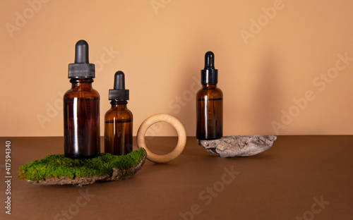 Serum bottles on brown with a beige background. Sustainable lifestyle concept  natural cosmetics. Cosmetic product decorated with moss  stones and wooden ring. Front view  copy space.