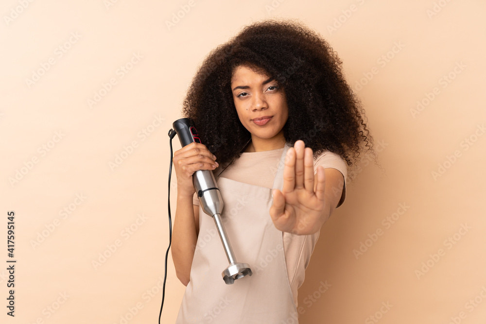 Young african american woman using hand blender isolated on beige background making stop gesture with her hand Young african american woman using hand blender isolated on beige background making stop gesture with her hand