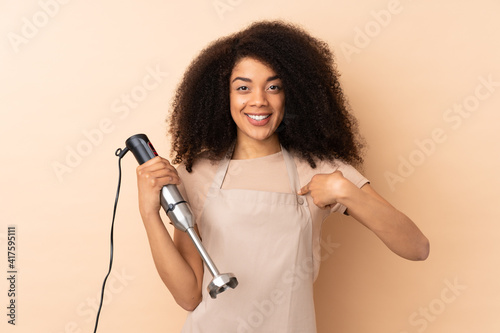 Young african american woman using hand blender isolated on beige background with surprise facial expression