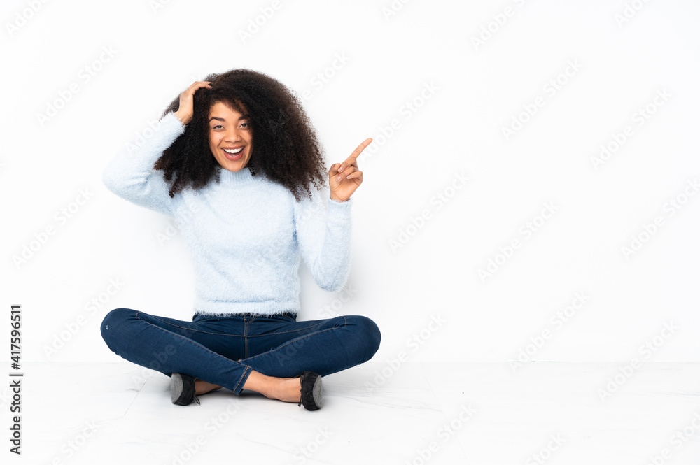 Young african american woman sitting on the floor surprised and pointing finger to the side Young african american woman sitting on the floor surprised and pointing finger to the side