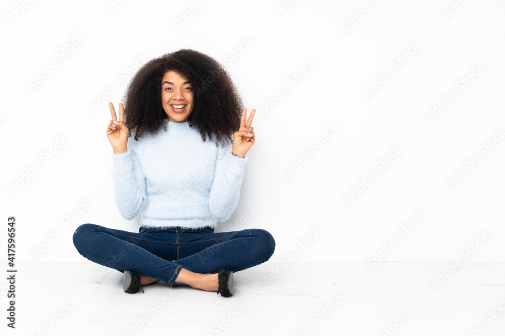 Young african american woman sitting on the floor showing victory sign with both hands Young african american woman sitting on the floor showing victory sign with both hands