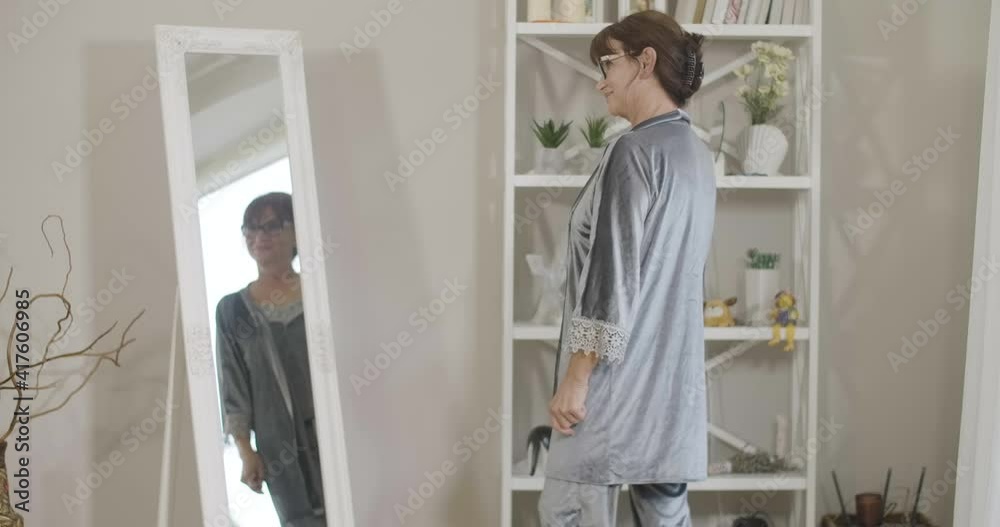 Side view of slim beautiful Caucasian senior woman in silk dressing gown looking at reflection in mirror and smiling. Confident gorgeous retiree admiring new clothes at home. Cinema 4k ProRes HQ. Side view of slim beautiful Caucasian senior woman in silk dressing gown looking at reflection in mirror and smiling. Confident gorgeous retiree admiring new clothes at home. Cinema 4k ProRes HQ.